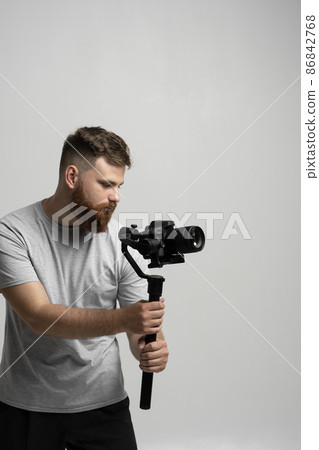 Professional male videographer filmmaker cinematographer dop shooting video using modern dslr camera on 3-axis gimbal over white wall. 86842768