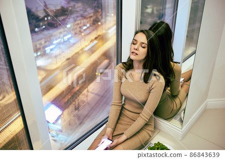 Floor to ceiling window with 20 years old woman with closed eyes and handy in hands. 86843639