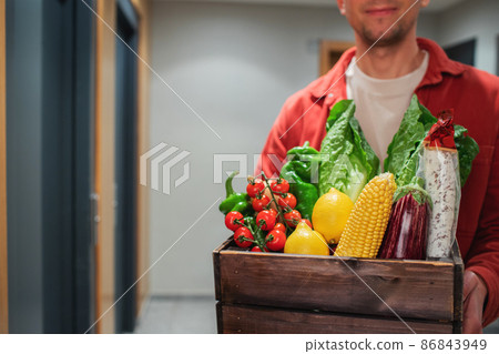 Delivery man holding paper bag with food in the entrance. The courier gives the box with fresh vegetables and fruits to the customer 86843949