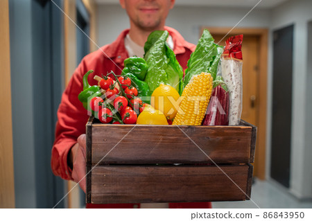 Delivery man holding paper bag with food in the entrance. The courier gives the box with fresh vegetables and fruits to the customer Delivery man holding paper bag with food in the entrance. The courier gives the box with fresh vegetables and fruits to the customer 86843950