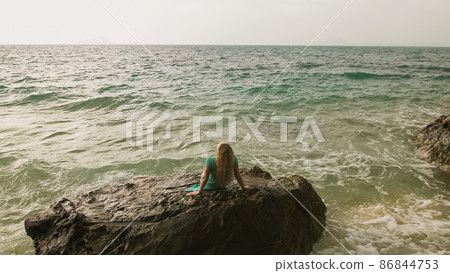 Woman sitting on rock of sea reef stone, stormy cloudy ocean. Blue swimsuit dress tunic. Concept resort coastline tourism summer holidays 86844753