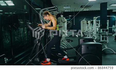 Fit Athletic Woman Exercising on an Elliptical Machine. Muscular Women Activity Training in Modern Gym. Workout in an Elliptic Walking Trainer. Fit Athletic Woman Exercising on an Elliptical Machine. Muscular Women Activity Training in Modern Gym. Workout in an Elliptic Walking Trainer. 86844759