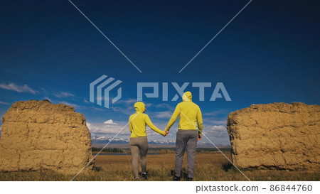 Man and woman in yellow green sportswear. Lovely couple of travelers hug and kiss near old stone enjoying highland landscape. Man and woman in yellow green sportswear. Lovely couple of travelers hug and kiss near old stone enjoying highland landscape. 86844760