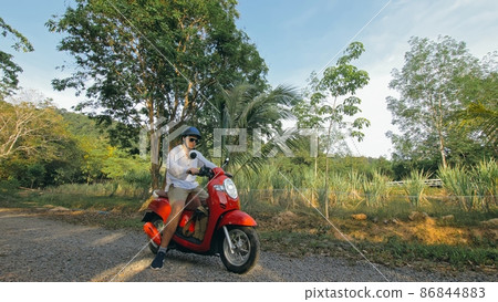Man on red motorbike in white clothes drive on forest road trail trip. One men caucasian tourist go on scooter, nearby tropical palm tree. Man on red motorbike in white clothes drive on forest road trail trip. One men caucasian tourist go on scooter, nearby tropical palm tree. 86844883