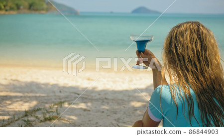 The woman is holding a glass of blue curacao cocktail, on beach azure blue sea of a tropical island. Blurred beautiful seaside background. The woman is holding a glass of blue curacao cocktail, on beach azure blue sea of a tropical island. Blurred beautiful seaside background. 86844903
