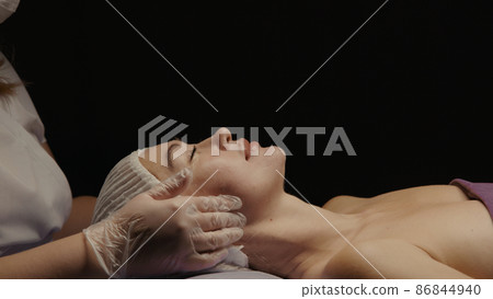 Woman receiving anti-ageing facial massage in spa salon relax. Wellness body skin care face beauty treatment. Black background Woman receiving anti-ageing facial massage in spa salon relax. Wellness body skin care face beauty treatment. Black background 86844940