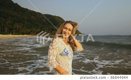 Woman is Wetting her Hair in a White Tunic on the Beach, near the Sea. Blonde in Sunglasses. Tourist Enjoying Vacation Time, Walks has Fun. Woman is Wetting her Hair in a White Tunic on the Beach, near the Sea. Blonde in Sunglasses. Tourist Enjoying Vacation Time, Walks has Fun. 86844944