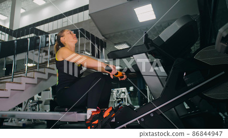 Fit Athletic Baby Woman Work out on Rowing Machine, Doing Her Fitness Exercise. Girl Activity Training in Modern Gym. Exercising on Row Machine. Fit Athletic Baby Woman Work out on Rowing Machine, Doing Her Fitness Exercise. Girl Activity Training in Modern Gym. Exercising on Row Machine. 86844947