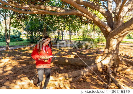 A charming little girl with long brown hair reads a book in the fresh air on a tree in an autumn park or in a forest glade. Girl who loves books. Education, reading, children's leisure A charming little girl with long brown hair reads a book in the fresh air on a tree in an autumn park or in a forest glade. Girl who loves books. Education, reading, children's leisure 86846379