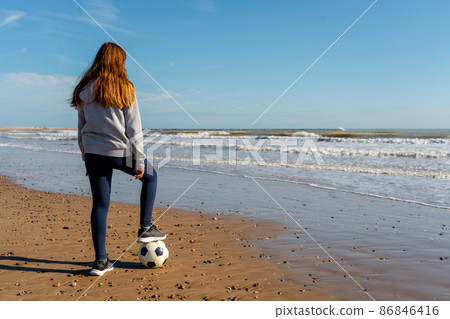Spanish pre-adolescent girl looking at the horizon on the shore of Burriana beach stepping on her soccer ball with one foot. Concept of illusion and hope to achieve your dreams. 86846416