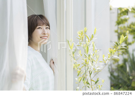 一個二十多歲的微笑女人的肖像,她拉開窗簾,向外看 一個二十多歲的微笑女人的肖像,她拉開窗簾,向外看 86847224