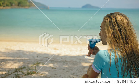 The woman is holding a glass of blue curacao cocktail, on beach azure blue sea of a tropical island. Blurred beautiful seaside background. The woman is holding a glass of blue curacao cocktail, on beach azure blue sea of a tropical island. Blurred beautiful seaside background. 86847499