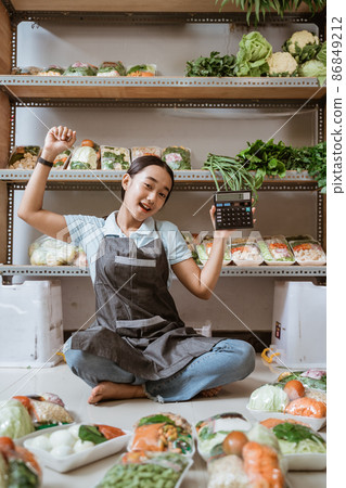 Excited veggie seller in apron sitting on floor using calculator 86849212