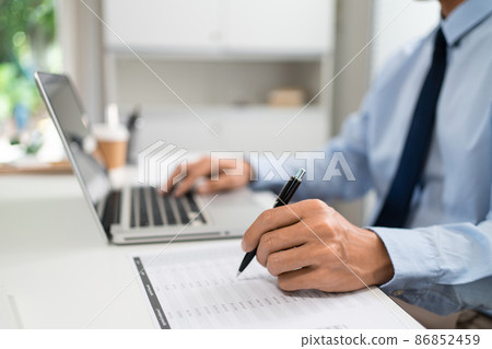 Working Man Conept The male officer sitting in the office, laying his left hand on the laptop, and writing something on the paper with his right hand Working Man Conept The male officer sitting in the office, laying his left hand on the laptop, and writing something on the paper with his right hand 86852459