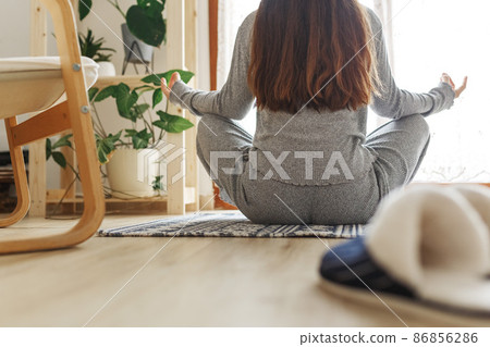 Young female sitting cross-legged in Easy pose, Sukhasana posture and meditating. Practicing yoga at home.Happy young woman enjoying sunny morning meditation routine.  86856286