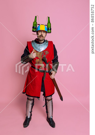 Full-length portrait of funny looking man, medieval warrior or knight in beer helmet and protective armor isolated over pink studio background 86858752