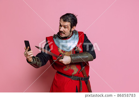 Comic portrait of medieval warrior or knight wearing wearing armor clothing using phone isolated over pink background. Comparison of eras, history 86858783