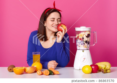 Beautiful woman makes a smoothie with blender in kitchen. Brunette girl likes tasty smell of apple and juice. Female with red hair band and blue shirt looks happy at home. Fruit and vegetables Beautiful woman makes a smoothie with blender in kitchen. Brunette girl likes tasty smell of apple and juice. Female with red hair band and blue shirt looks happy at home. Fruit and vegetables 86863570
