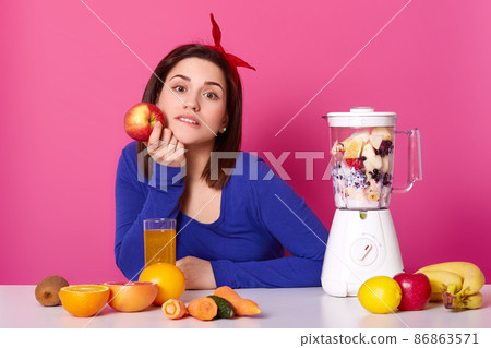 Cute dark haired thoughtful girl with red hair band and blue shirt holds apple in hand. Woman makes fruit smoothie. Lady uses blender at home. Carrots on table. Housewife isolated over pink Cute dark haired thoughtful girl with red hair band and blue shirt holds apple in hand. Woman makes fruit smoothie. Lady uses blender at home. Carrots on table. Housewife isolated over pink 86863571