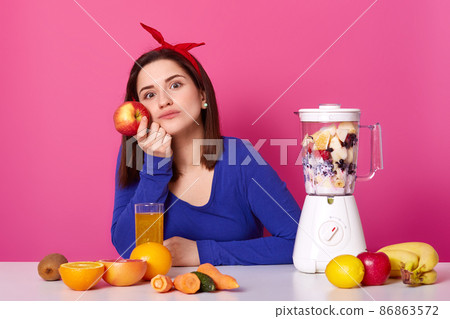Beautiful young girl wears blue sweater and with red hairband sits at table and thinks about her plans for evening. Lady holds fresh apple in hand. Charming woman full of thoughts and dreams. Beautiful young girl wears blue sweater and with red hairband sits at table and thinks about her plans for evening. Lady holds fresh apple in hand. Charming woman full of thoughts and dreams. 86863572