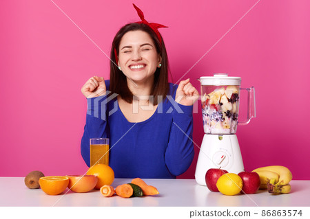 Horizontal shot of cheerful young woman makes smoothie with blender. Beautiful brunette with bended elbows and closed eyes looks happy while preparing tasty fruit cocktail. Healhy lifestyle concept. Horizontal shot of cheerful young woman makes smoothie with blender. Beautiful brunette with bended elbows and closed eyes looks happy while preparing tasty fruit cocktail. Healhy lifestyle concept. 86863574