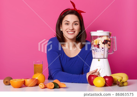 Positive woman with cheerful facial expression, being in high spirit, mixes fruit in blender for making smoothie, dressed in blue jumper and hair band, likes to drink fresh juice. Lifestyle concept. Positive woman with cheerful facial expression, being in high spirit, mixes fruit in blender for making smoothie, dressed in blue jumper and hair band, likes to drink fresh juice. Lifestyle concept. 86863575