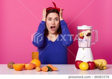 Surprised beautiful woman wears blue sweater and headband, keeps hands on head, has shocked expression, opens mouth from astonishment, surrounded with fruit, vegetables and blender, makes smoothie. Surprised beautiful woman wears blue sweater and headband, keeps hands on head, has shocked expression, opens mouth from astonishment, surrounded with fruit, vegetables and blender, makes smoothie. 86863576