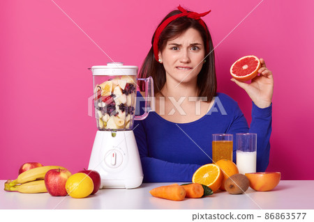 Confused black haired emotional female sits at table, biting her lip, holding half of grapefruit in one hand, mixing fruit in blender, making sweet nutritious smoothie for healthy meal on diet. Confused black haired emotional female sits at table, biting her lip, holding half of grapefruit in one hand, mixing fruit in blender, making sweet nutritious smoothie for healthy meal on diet. 86863577