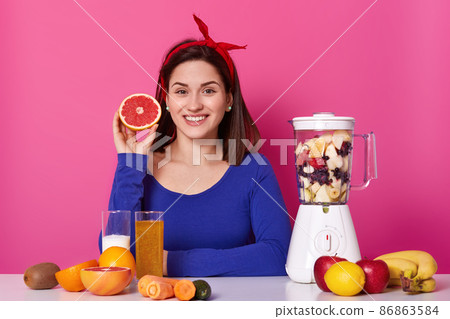 Portrait of smiling positive female holding cut fruit in her hand. Young model waits for smoothie to be ready. Cheerful happy woman stick to healthy diet with pleasure. Healthy lifestyle concept. Portrait of smiling positive female holding cut fruit in her hand. Young model waits for smoothie to be ready. Cheerful happy woman stick to healthy diet with pleasure. Healthy lifestyle concept. 86863584