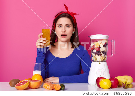 Close up of brunette woman with glass of fresh orange juice in hand. Lady likes healthy eating, making smoothie at home. Surprised female with blender against pink wall. Dieting and people concept. Close up of brunette woman with glass of fresh orange juice in hand. Lady likes healthy eating, making smoothie at home. Surprised female with blender against pink wall. Dieting and people concept. 86863586