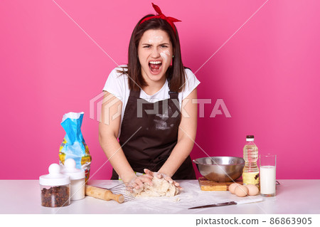 Beautiful angry cook kneads dough and screams loud, being sick and tired of preparing homemade pastry. Furious brunette woman works in kitchen and dreams of having rest. Culinary and food concept. Beautiful angry cook kneads dough and screams loud, being sick and tired of preparing homemade pastry. Furious brunette woman works in kitchen and dreams of having rest. Culinary and food concept. 86863905