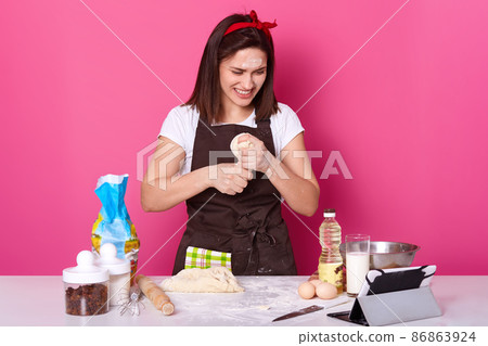 Happy housewife wears kitchen apron dirty with flour, white t shirt, red headband, kneads dough while having videocall with her husband. Studio picture of smiling baker isolated over pink background. 86863924