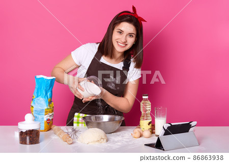 Image of woman making and preparing pastry in bakery kitchen. Adding flour bits. Female has pleasant facial expession, looks happilly directly at camera, baking bread, dressed brown apron and t shirt. Image of woman making and preparing pastry in bakery kitchen. Adding flour bits. Female has pleasant facial expession, looks happilly directly at camera, baking bread, dressed brown apron and t shirt. 86863938
