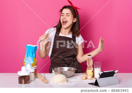 Photo of beautiful woman making cake. Lady stands over rose wall, holds whisk as microphone and sings songs, listening music while baking pie, dresses t shirt, apron and has hair band. Copy space. 86863941