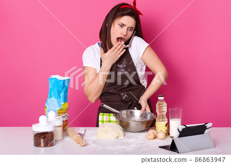 Impressed surprised emotional model poses isolated over pink background, wearing brown apron and white t shirt , talking over her mobile phone, looks shocked by news, cooking dough. Baking concept. 86863947