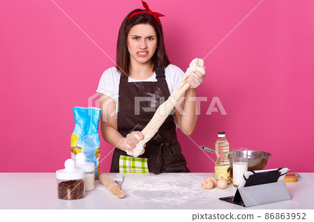 Dissatisfied woman kneading dough, baking cake or pie, housewife bakes at home, brunette lady surraunded products, dislikes to spend time in kitchen, making homemade pastry, posing on pink background. 86863952