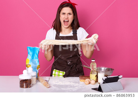Emotional expressive woman in kitchen, shouting female surrounded with tools for cooking food, lady kneads dough with angry facial exxpression, young girl wearing white casual t shirt and brown apron. Emotional expressive woman in kitchen, shouting female surrounded with tools for cooking food, lady kneads dough with angry facial exxpression, young girl wearing white casual t shirt and brown apron. 86863953