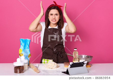 Horizontal shot of angry housewife, lady wearing brown apron dirty with flour, white casual t shirt, red headband, keeps hands up, looks tired, model posing isolated over pink studio background. 86863954