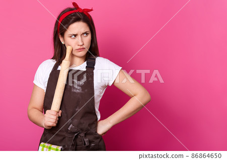 Brunette cook with red hair band, brown apron soiled with flour and white t shirt decides what recipe to use for baking pie. Young baker holds rolling pin and touches cheek with it. Culinary concept. 86864650
