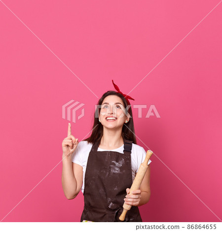 Image of smiling female chef, cook or baker with rolling pin in hands pointing up with her index finger and looking up, isolated over pink background, copy space for advertisment or promotional text. Image of smiling female chef, cook or baker with rolling pin in hands pointing up with her index finger and looking up, isolated over pink background, copy space for advertisment or promotional text. 86864655