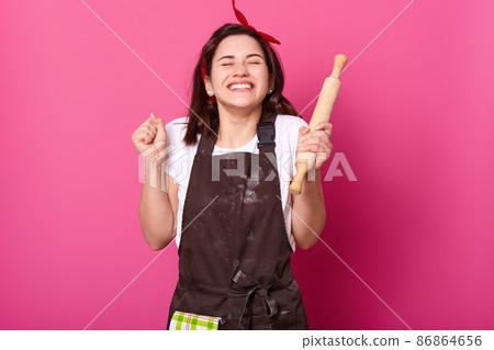 Baker woman holds baking rolling pin, wears brown apron, white t shirt. Young girl bends elbows and shouts yes with closed eyes, female maskes cake first time. Cook isolated on pink background. Baker woman holds baking rolling pin, wears brown apron, white t shirt. Young girl bends elbows and shouts yes with closed eyes, female maskes cake first time. Cook isolated on pink background. 86864656