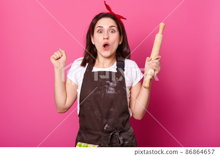 Young baker against pink wall looks at camera, bends elbows, holds rolling pin. Charming lady with happy facial expression has new idea while baking chocolate cookies. Food and culinary concept. Young baker against pink wall looks at camera, bends elbows, holds rolling pin. Charming lady with happy facial expression has new idea while baking chocolate cookies. Food and culinary concept. 86864657