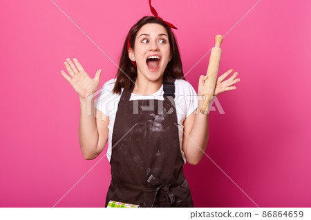 Attractive emotional young beauty screaming with delight, raising her hands, looking aside, opening mouth widely, having wooden rolling pin in hand, looks inspired, making out new culinary recipe. Attractive emotional young beauty screaming with delight, raising her hands, looking aside, opening mouth widely, having wooden rolling pin in hand, looks inspired, making out new culinary recipe. 86864659