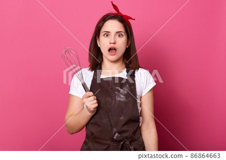 Image of astonished good looking female standing isolated over pink background in studio, opening her eyes and mouth widely, looking directly at camera, holding whisk in one hand. Cook concept. Image of astonished good looking female standing isolated over pink background in studio, opening her eyes and mouth widely, looking directly at camera, holding whisk in one hand. Cook concept. 86864663