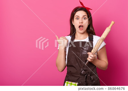 Portrait of young attractive woman in brown apron poses in studio, points thumb aside isolated on rose background. Astonished female stands with opened mouth, holds rolling pin. Culinary concept. 86864676
