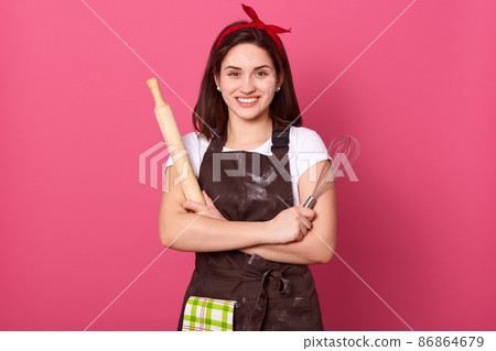 Horizontal shot of confident cheerful beautiful young woman posing isolated over pink background in studio, having folded arms, holding rolling pin and whisk in both hands. Culinary concept. 86864679