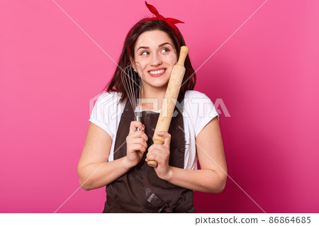 Cheerful smiling young female poses isolated over pink background, touching face with whisk and wooden rolling pin, looing aside. Slender energetic model wears brown apron, t shirt and red headband. Cheerful smiling young female poses isolated over pink background, touching face with whisk and wooden rolling pin, looing aside. Slender energetic model wears brown apron, t shirt and red headband. 86864685