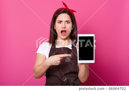 Portrait of surprised dark haired young housewife recieves strange message on phone, stares with unexpected gaze at camera and keeps mouth opened, dressed in white t shirt, brown apron, red hair band. Portrait of surprised dark haired young housewife recieves strange message on phone, stares with unexpected gaze at camera and keeps mouth opened, dressed in white t shirt, brown apron, red hair band. 86864702