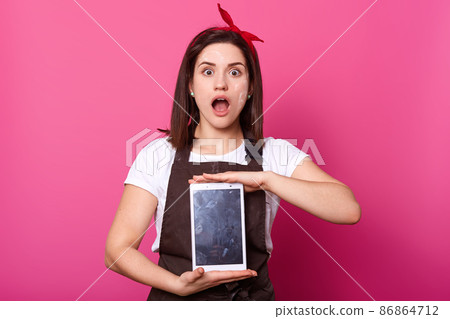 Studio shot of woman wears brown apron, showing blank empty screen with copy space. Caucasian model has astonished facial expression, holds digital gadget in her hand, poses with open mouth. Studio shot of woman wears brown apron, showing blank empty screen with copy space. Caucasian model has astonished facial expression, holds digital gadget in her hand, poses with open mouth. 86864712