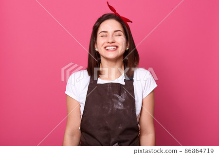 Indoor shot of beautiful magnetic cute young woman standing isolated over pink background in studio, closing eyes, smiling sincerely, wearing brown dirty apron, white t shirt and red headband. 86864719